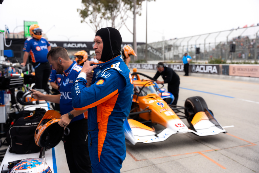 Scott Dixon puts on his racing equipment while his pit crew prepares his car in the background.