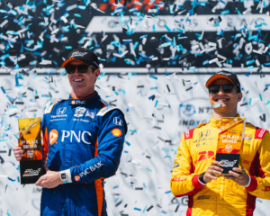Scott Dixon holds up a first-place trophy after winning a race as confetti falls around him.