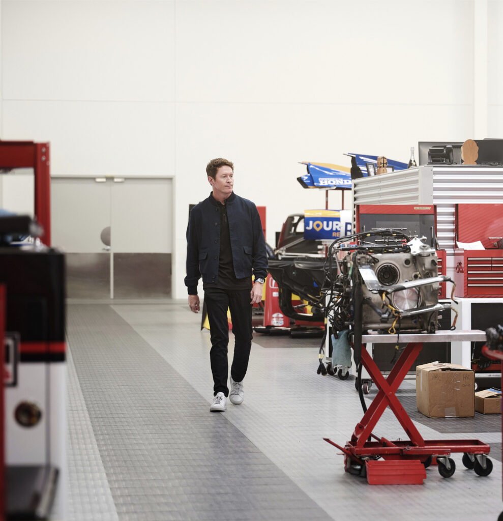 Scott Dixon walks through an auto shop, passing various cars and car parts.